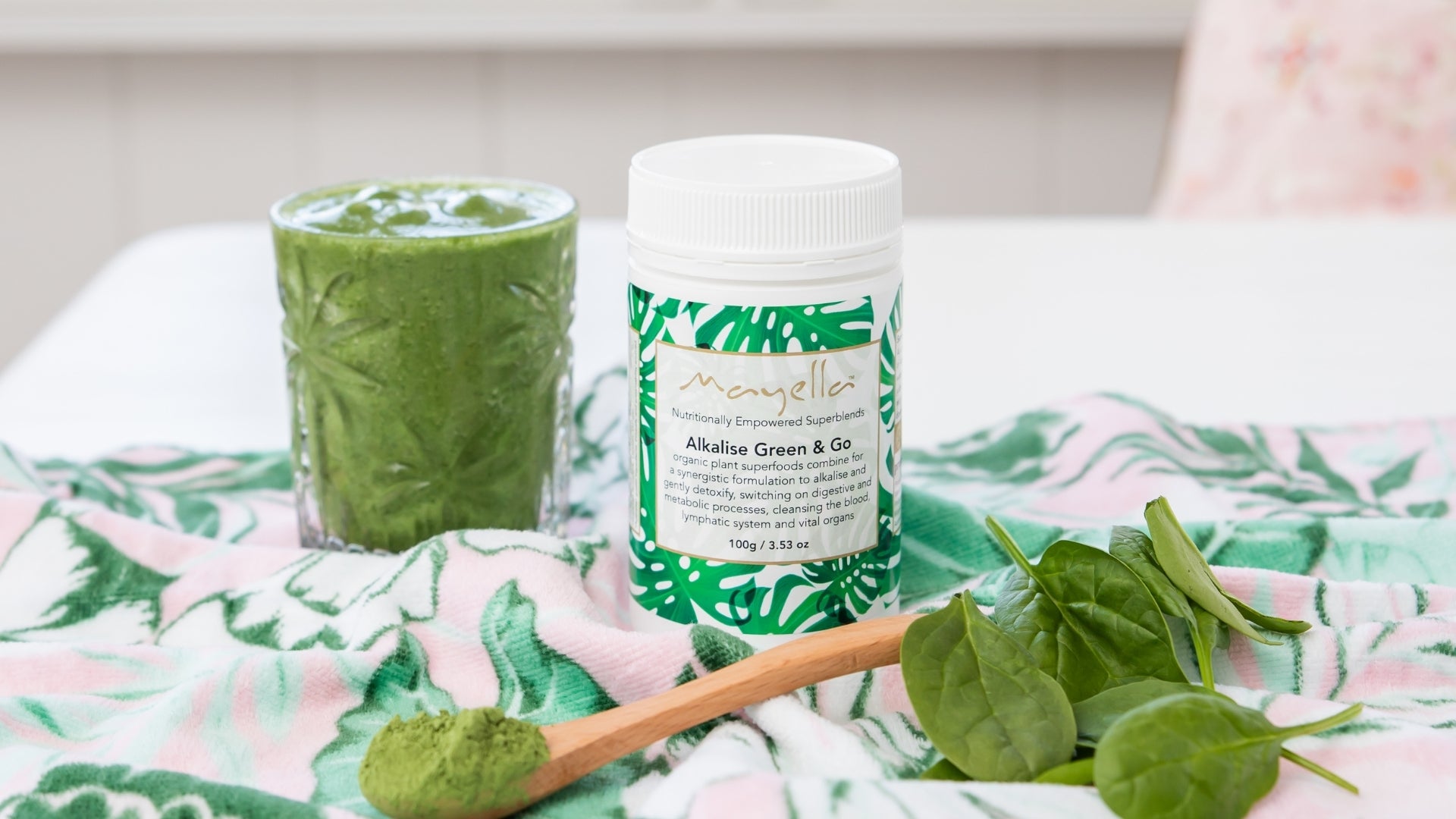 Alkalise Green & Go tub next to a green smoothie sitting on a tropical towel surrounded by spinach and greens powder