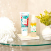 tube of AHA exfoliating whip standing next to AHA efoliating serum on a stone tile on a glass shelf in the shower next to a body scrubber and a small white vase of yellow flowers
