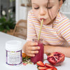 young girl drinking a deep purple smoothie out of a small glass milk jar next to half a pomegranate and sliced strawberries