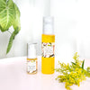 two cleansing serum bottles side by side in 15ml and 100ml sizes on a white table with green leaves on the left hand side of the photo and yellow flowers on the right hand side