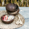 stack of three empty low-set coconut bowls on a flax place mat next to a coconut bowl filled with an acai smoothie topped with coconut and strawberries
