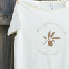 close up of a creme-coloured t-shirt hanging against a timber fence, the design on the shirt is a gold leaf with text circling it that reads you gotta nourish to flourish
