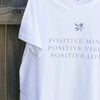 close up of a white t-shirt hanging against a timber fence, the design on the shirt is a small silver leaf with silver text underneath that reads positive mind, positive vibes, positive life