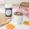 white tea cup holding a metal tea strainer filled with rest easy tisane on a white table top next to a container of rest easy, a woven place mat and two dehydrated orange slices