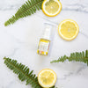 flat lay image of a bottle of vitamin c lift serum on a white  and grey marble bench top next to three slices of lemon and three fern fronds