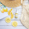 flat lay image of a bottle of vitamin c lift serum on a white timber table next to slices of lemon, a woven shopping bag, a beach hat and a magazine