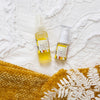 birds eye view of two vitmain c lift serums on a white textured bed cover next to a yellow/orange fabric