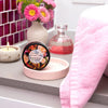 round container of milk and honey powder foundation standing on its rounded side on a pink stone coaster on a grey bathroom vanity with a candle, flowers, perfume and bold purple tiles in the background