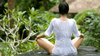 The back of a woman in a seated meditation position facing a garden of greenery. She is wearing a white, slightly see-through tunic and has short black hair.