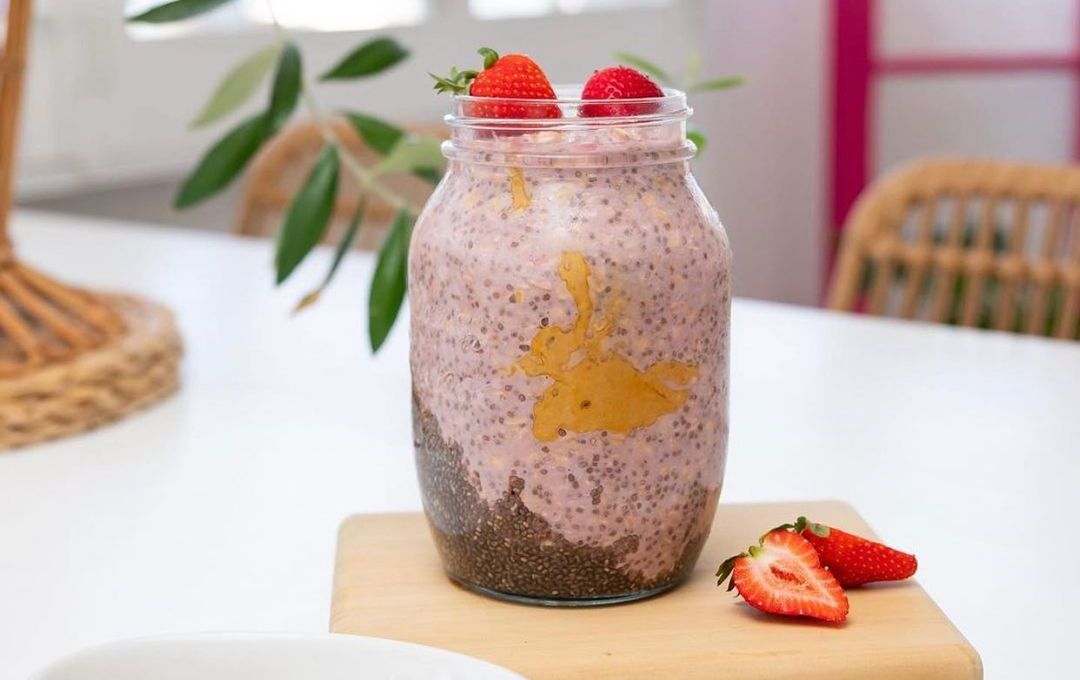 A large 1L jar filled with pink-colours chia and oat porridge. The jar is on a small timber chopping board with two strawberry halves next to it. The chopping board is on a white dining room table.