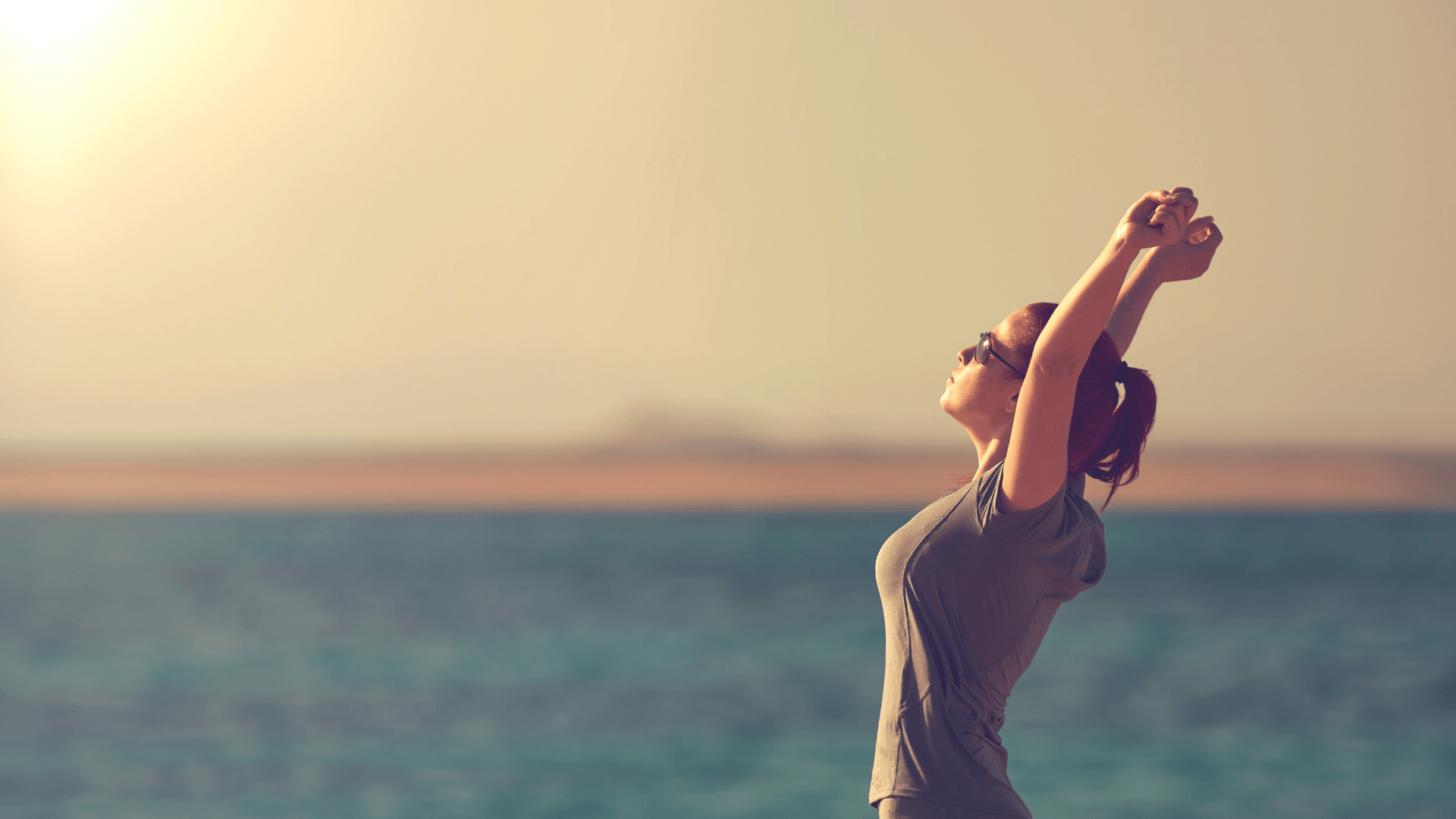 A photo of a woman, side-on, standing with her arms raised above her head. She is facing the sun to the left of the photo and looks like she has taken a deep breath in, chest open, back arched.