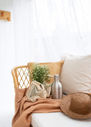 Mayella Stainless Steel 1.9L drinking flask sitting on a beige cord throw rug next to a white mesh shopping tote with fresh wild flowers on cane couch with a straw hat and white cotton pillow.