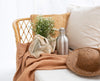 Mayella Stainless Steel drinking flask sits on a throw blanket on a cane couch. A string bag with daisy flowers and a woven hat sit next to the steel flask. White and a straw cushion frame the flask.  