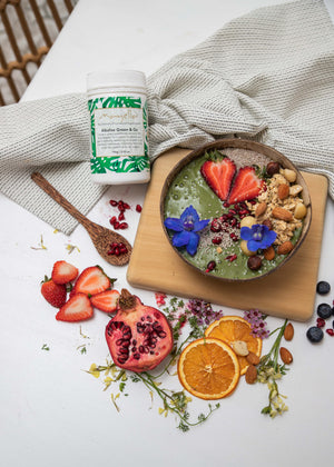 Mayella Alkalise Green & Go lying on a grey and white cotton tea towel next to a green smoothie bowl topped with fresh fruits, nuts  seeds and blue edible flowers surrounded b slices of pomegranate, strawberries , sprigs of flowers and oranges.