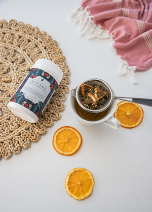 Mayella organic loose leaf tisane Rest Easy lying on a straw mat next to a white china cup filled with a fresh cup of Rest easy tea. The mesh strainer with rest easy tea sitting in the cup and slices of orange on the table