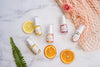 Five of Mayella natural and organic vegan skincare 15ml sizes lying across a grey and white marble table with apricot mesh shopping tote, slices of orange and pink rose.