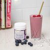 tub of acai berry beautiful on a white stone kitchen bench next to a glass full of pink smoothie with two cookbooks in the background