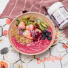 bright pink smoothie bowl topped with buckwheat, sliced strawberries and kiwi, fresh blueberries and pomegranite seeds next to a tub of acai berry beautiful