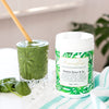 tub of alkalise green and go next to a glass filled with green smoothie on a white table  surrounded by ice and spinach leaves