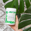a woman's hand holding a tub of alkalise green and go next to two leaves of a monstera plant