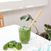 a green smoothie in a short glass on a white table with a bamboo straw in it and baby spinach leaves next to it