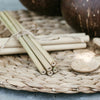 two bunches of bamboo straws both tied together with twine resting on a woven place mat next to a wooden spoon and two coconut bowls