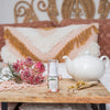 bottle of cleansing serum on a timber tray next to a tea pot and pink wild flowers all resting on a bed with boho style cushions in the background