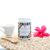 tub of ginger kisses tisane next to a white teacup, a pink hibiscus flower and a wooden spoon all on a woven place mat