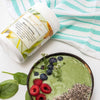 a green smoothie bown topped with raspberries, blueberries and chia seeds next to a tub of green harmony blend on a white table top