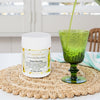 tub of green harmony capsules on a woven placemat next to a vibrant green glass with a green juice being poured in