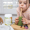 young girl drinking a green smoothie out of a small glass milk jar with a container of green harmony to her right and some blueberries and nuts to her left