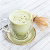 light green matcha latte in a clear glass mug on an earthy green and and brown saucer next to a matcha chasen or whisk