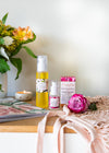 Table with vase of orange, white, yellow flowers, a small candle in a pottery dish, wellness magazine. Lying on the table is a pale peach string eco shopping tote with a beautiful pink rose bloom sitting on it. In the middle of these items is a 100ml bottle of Mayella Cleansing Serum and 1 15 ml Mayella Olivane Rose glass bottle with the Olivane Rose labelled box.