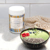 tub of nourish formulated blend on a white and grey stone bench top next to a coconut shell bowl filled with a green smoothie bowl topped with chia seeds, raspberries and blueberries