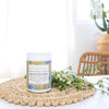 tub of nourish formulated blend on a woven place mat next to a bunch of small flowers with a cactus and white sheer curtain in the background