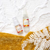 birds eye view of two mayella olivane repair serums citrus infusion in 15ml and 30 ml sizes laying on a textured white bed sheet next to a mustard coloured fabric throw