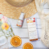 bottle of olivane repair serum citrus infusion on a white timber bench next to two slices of dehydrated orange, a cream netter shopping bag, a straw beach hat and an open magazine