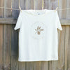 a creme-coloured t-shirt hanging against a timber fence, the design on the shirt is a gold leaf with text circling it that reads you gotta nourish to flourish