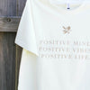 close up of a creme-coloured t-shirt hanging against a timber fence, the design on the shirt is a small gold leaf with gold text underneath that reads positive mind, positive vibes, positive life
