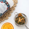 close up of dry loose rest easy tisane sitting in a tea strainer on a white benchtop next to a slice of orange and a container of rest easy tisane