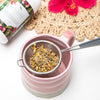 dry loose tisane of skin tonic sitting in a tea strainer rested on a big pink mug with a skin tonic tub, woven table mat and red flower in the background