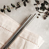 two metal straws side by side resting on a creme coloured cloth napkin on a grey marble bench top and next to four branches of with small deep green leaves