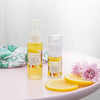 two bottles of vitamin c lift serum in 15ml and 30 ml sizes on a white glass stand next to two slices of lemon, two violet flowers and a green and pink beach towel in the background