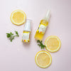 flay lay of two bottle of Vitamin C lift serum in two sizes next to three slices of lemon and on a light pink background