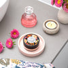 round container of vanilla cream powder foundation on a stone pink coaster sitting on a grey bathroom vanity next to a candle, three pink flowers and a small glass jar of pink perfume
