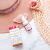 bottle of olivane repair serum resting on a pink rose quartz on a pink beach hat and next to its box and a netted pink shopping bag