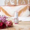 bottle of olivane repair serum rose infusion on a timber stand sitting on a bed with a boho style cushion in the background and pink, red and purple flowers next to it